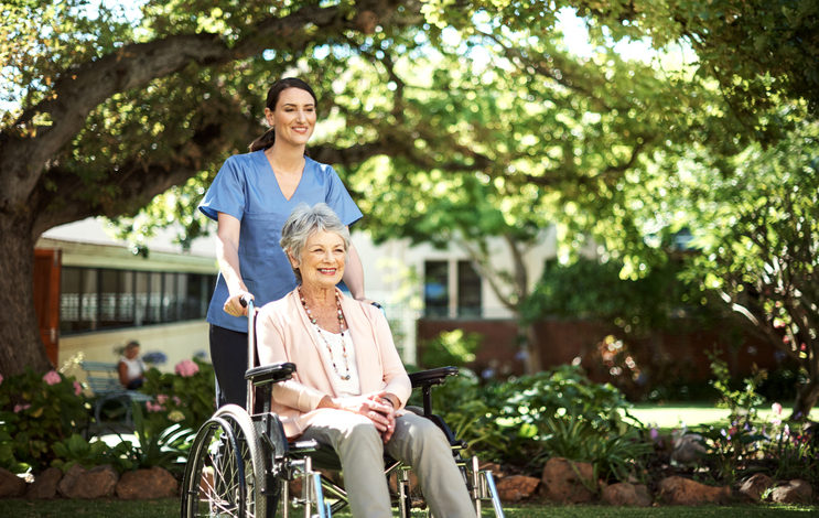 Nurse in scrubs pushes mature adult in pink top and necklaces in wheelchair under shaded area with trees