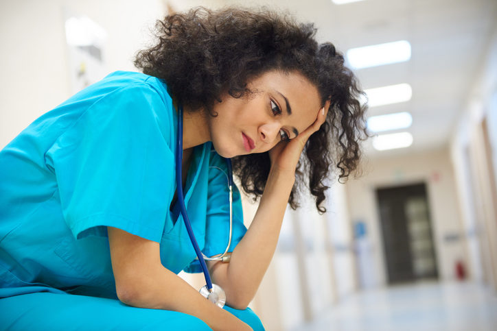 Seated nurse with curly dark hair rests head in hands, looks down