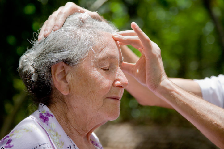 Senior woman sits outside while person's hand is massaging her forehead in front of background of trees