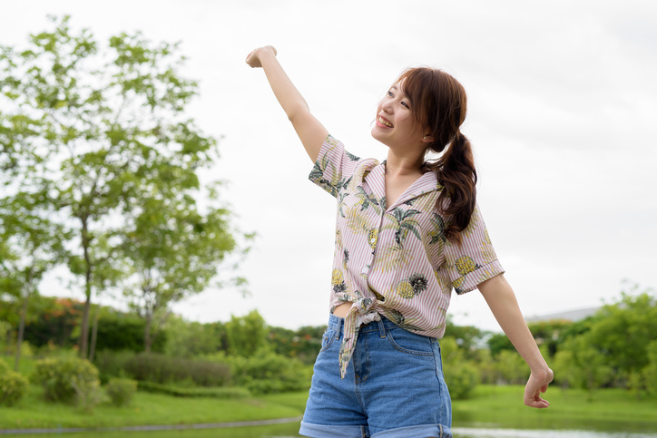 Young person in shorts with ponytail raises arms and does stretch outside near trees